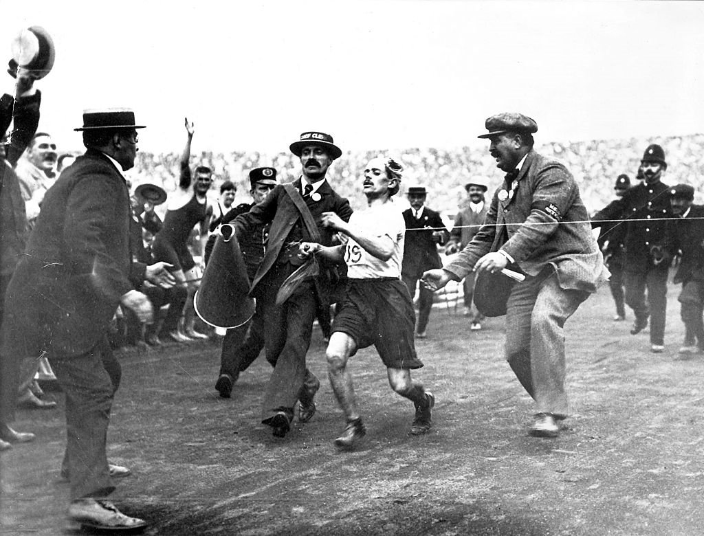 Photographie ancienne : un coureur épuisé soutenu par des officiels à l'arrivée du marathon dans un stade