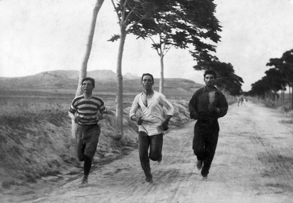 Photographie ancienne : trois coureurs sur une route de terre bordée d'arbres, marathon d'Athènes 1896