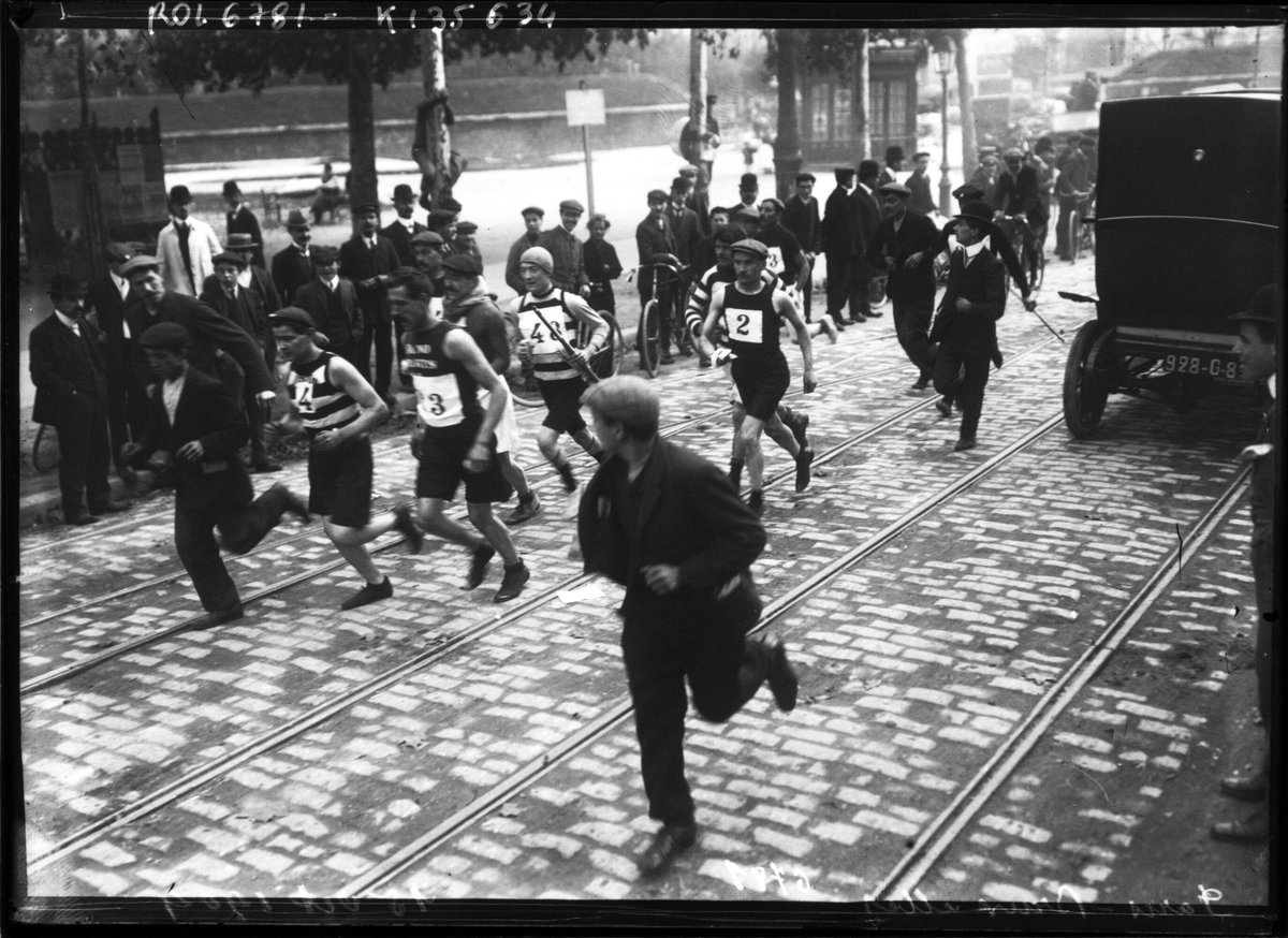 Photographie ancienne : des coureurs en maillot s'élancent dans une rue pavée, entourés de spectateurs