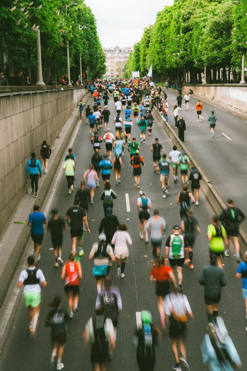 Peloton serré de coureurs vu de haut, sur une large route bordée d'arbres